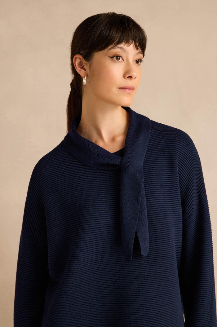 A woman with dark hair in a ponytail wears the Tie Neck Navy Ribbed Knit, standing against a beige background.