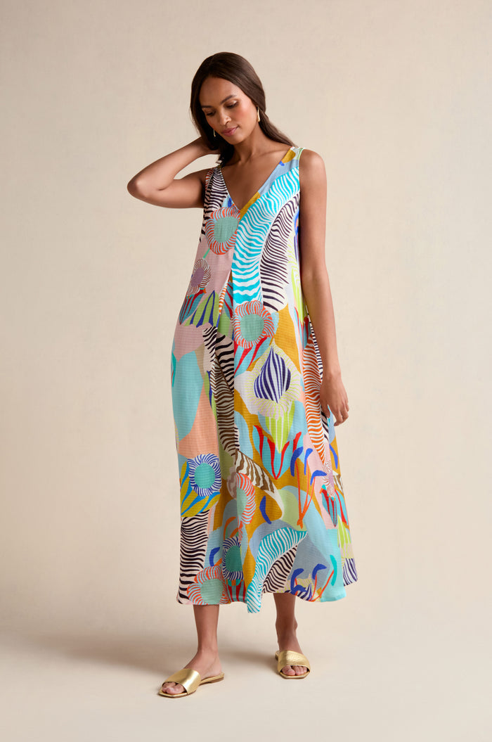 A woman models the Tropical Fantasy Sleeveless Dress, featuring a colorful abstract pattern, as she stands against a beige background with one hand in her hair.