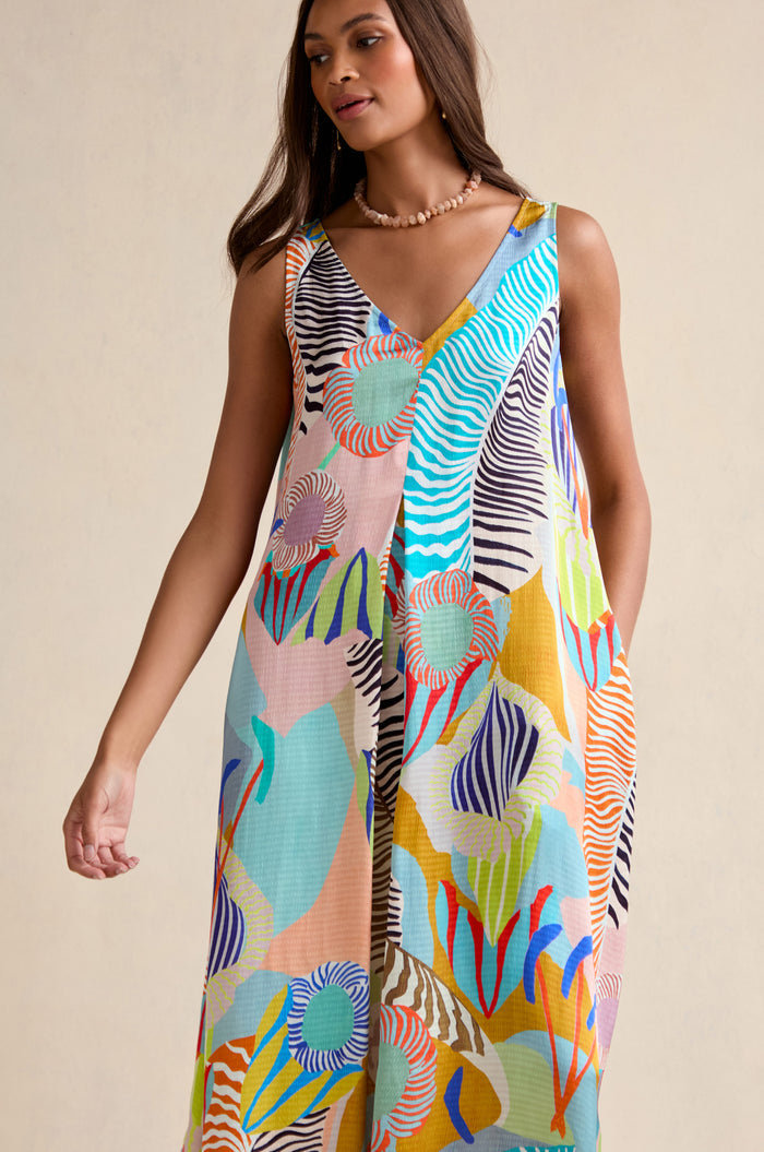 A woman models the Tropical Fantasy Sleeveless Dress, featuring a vibrant abstract pattern, against a neutral backdrop.