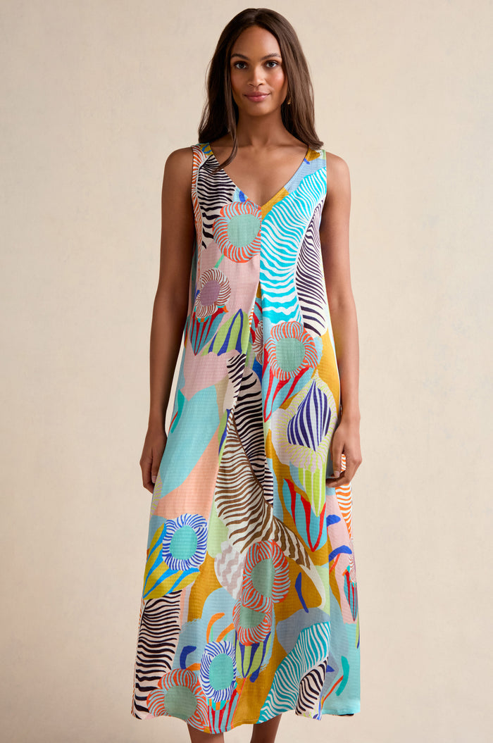 A woman stands against a plain background wearing the Tropical Fantasy Sleeveless Dress, featuring a colorful abstract pattern in blue, orange, green, and yellow tones.