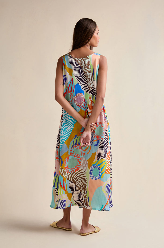 A woman faces away in a Tropical Fantasy Sleeveless Dress, an ankle-length maxi with a vibrant abstract print. She clasps her hands behind her back and wears flat yellow shoes.