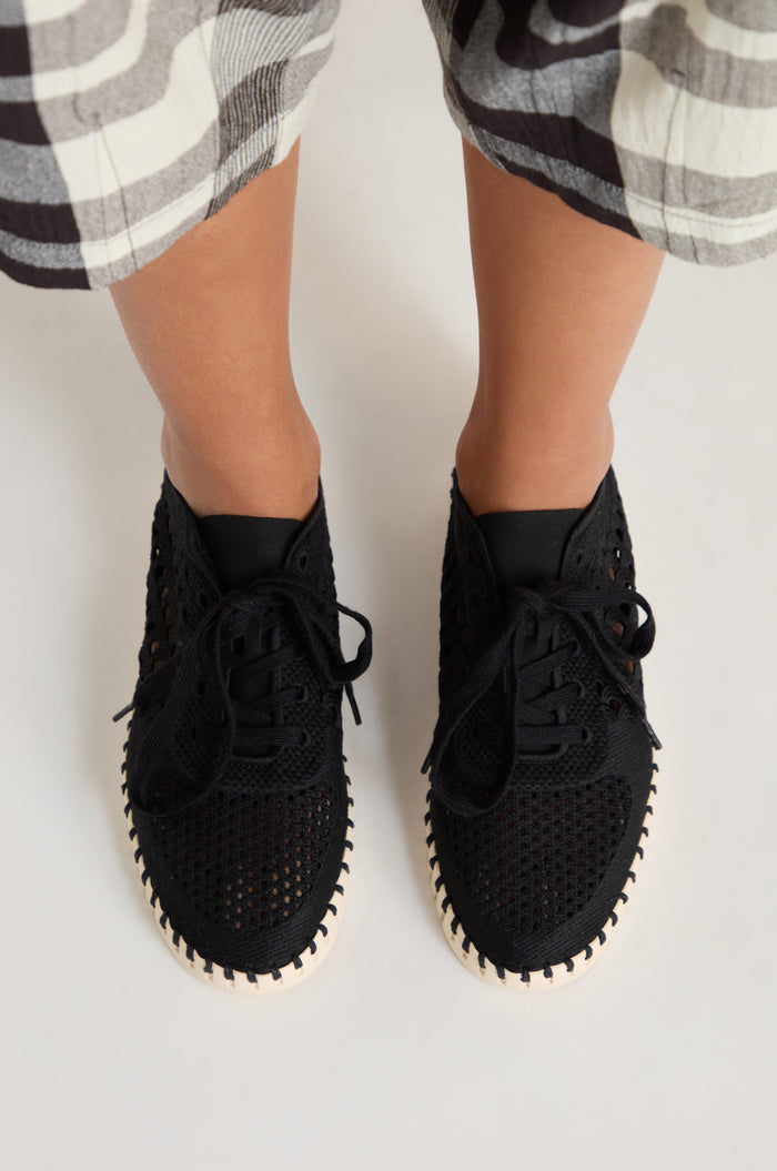 Person wearing black Tulip Embroidered Lace Up Trainers and black-and-white plaid pants, standing on a light-colored surface viewed from above.