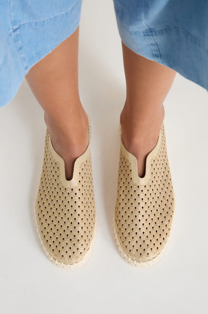 A person wearing Tulip Glitter Slip on Shoes with a tan perforated design stands on a white surface, with a blue garment visible above the knees.