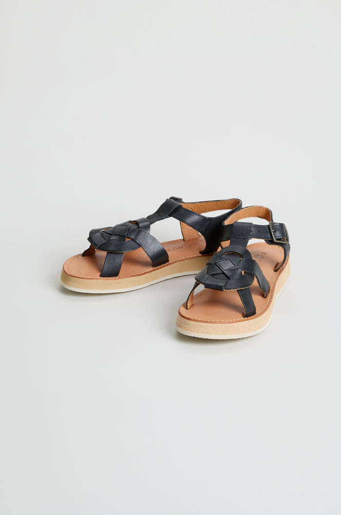 The Tulum Leather Sandals feature black leather woven straps, a buckle, and tan soles. Handcrafted by Mapache Tulum, they are shown on a plain white background.