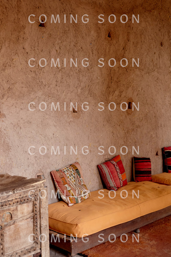 A rustic room with a textured plaster wall, wooden cabinet, and low seating with patterned cushions—perfect for lounging in the Cotton Jersey Boxy Pocket Top.