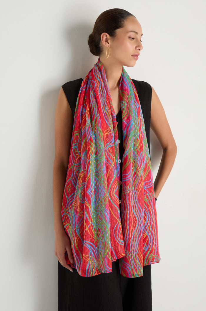 A woman in a sleeveless black dress stands against a white wall, wearing the Twirl Print Dobby Scarf draped around her neck and shoulders.