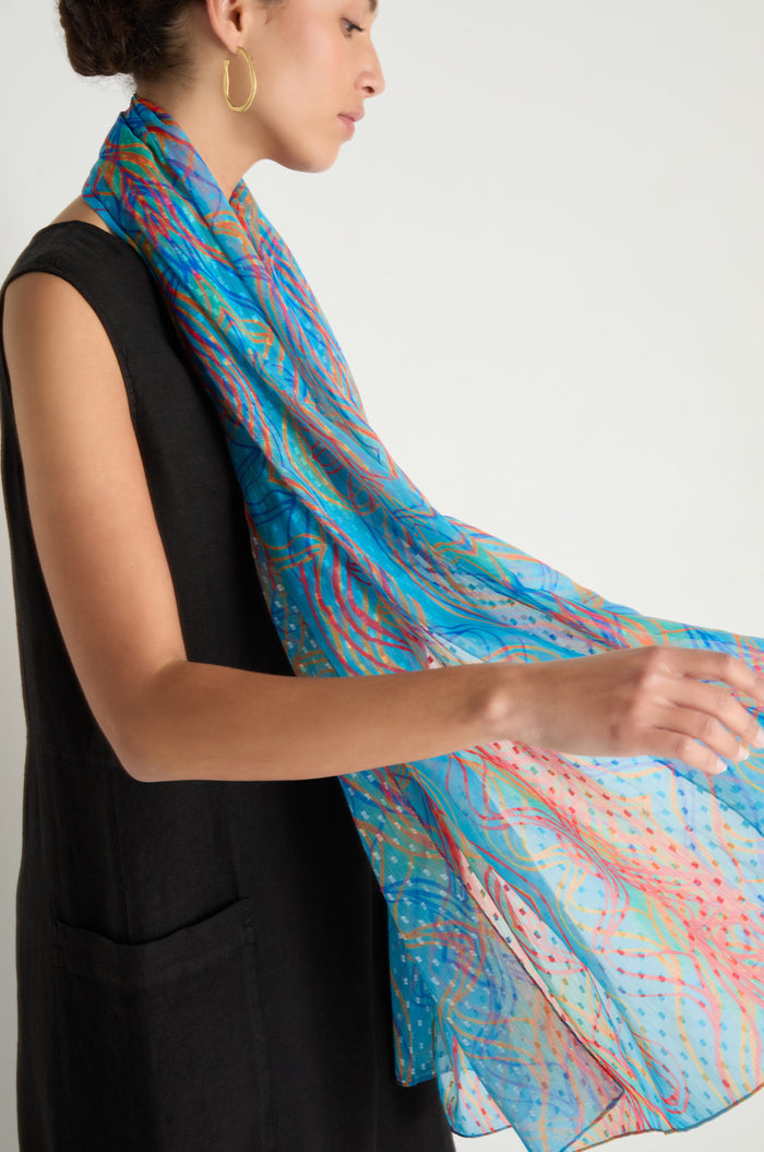 A woman in a sleeveless black dress and gold hoop earring holds the Twirl Print Dobby Scarf, which features lightweight fabric with multicolored wavy patterns and an abstract swirl print.