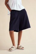 A person stands with one hand in a pocket, wearing a white short-sleeve top, Twisted Linen Wide Shorts in navy, and light-colored slip-on sandals.