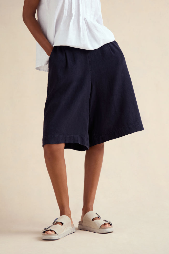 A person stands with one hand in a pocket, wearing a white short-sleeve top, Twisted Linen Wide Shorts in navy, and light-colored slip-on sandals.