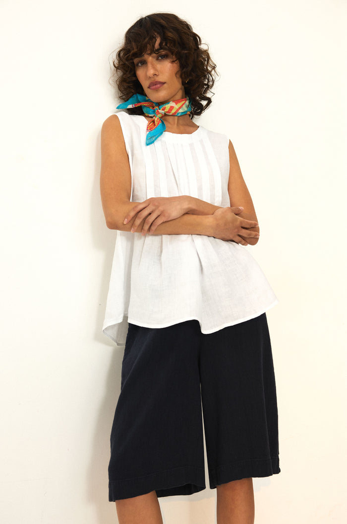 A woman stands against a white wall with crossed arms, wearing the Twisted Linen Wide Shorts featuring an elasticated waist, paired with a sleeveless white top and a colorful scarf around her neck.