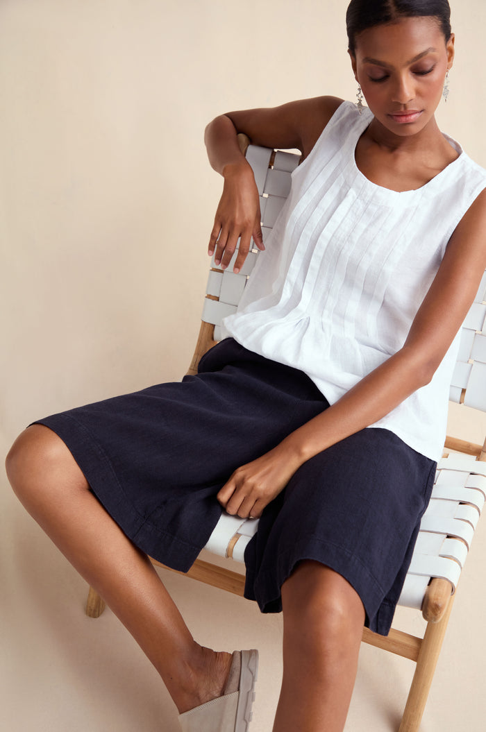 A woman sits on a woven chair wearing Twisted Linen Wide Shorts with a sleeveless white blouse and light slip-on shoes, set against a plain background.
