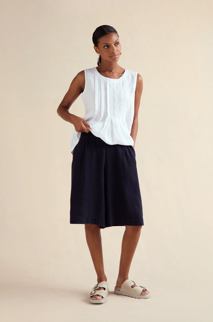 A woman stands against a plain background wearing a sleeveless white top, Twisted Linen Wide Shorts in black, and light-colored sandals.