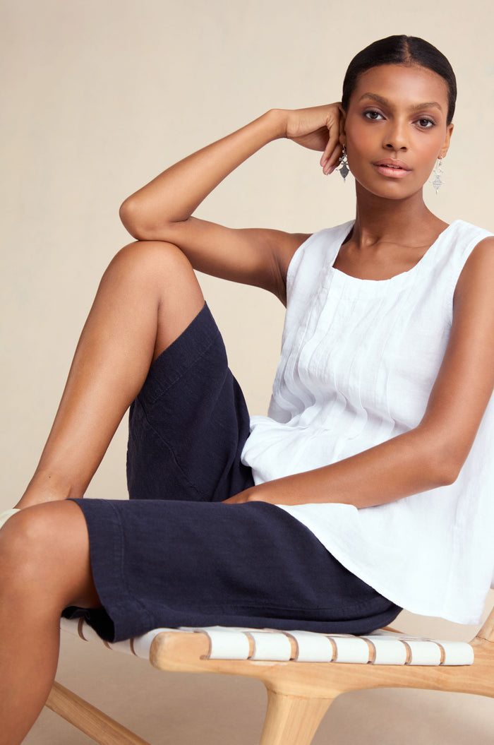 A woman with her hair pulled back sits on a chair, wearing a white sleeveless blouse and Twisted Linen Wide Shorts in navy, one arm resting on the chair.