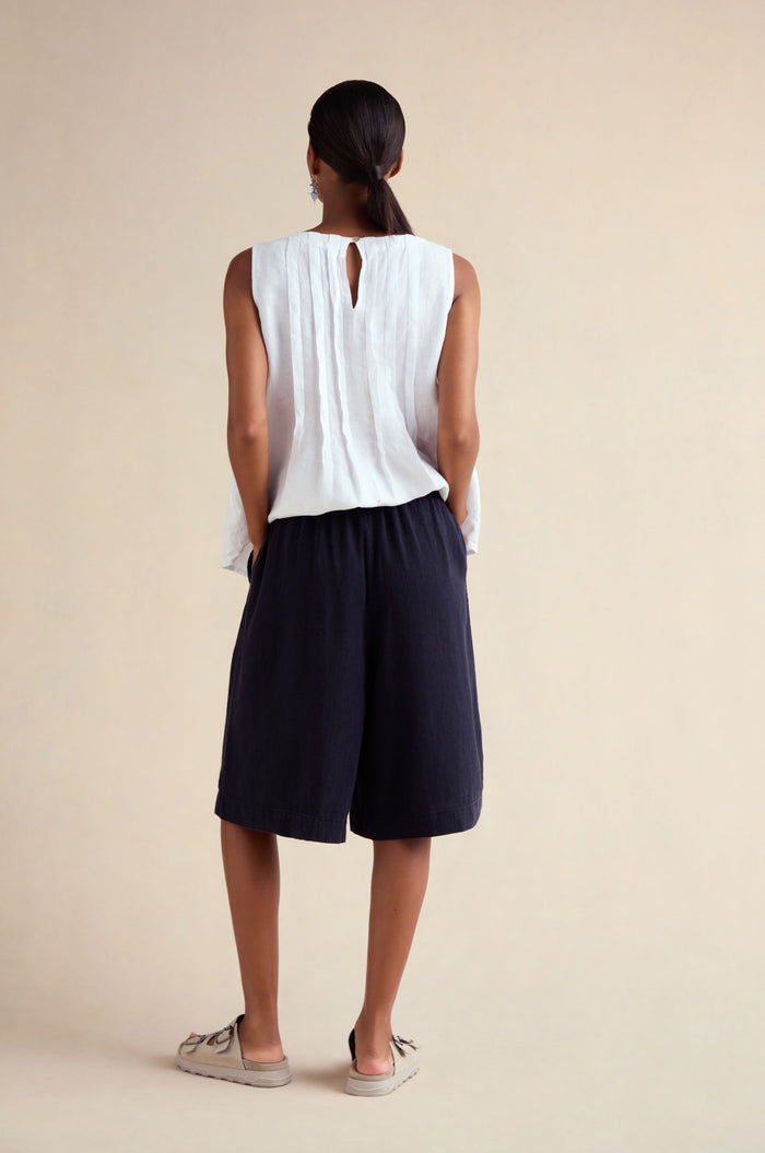 A woman stands with her back to the camera, wearing a sleeveless white top and Twisted Linen Wide Shorts, paired with beige flats, against a plain light background.