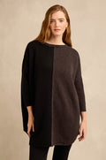 A woman wears the Two Colour Knit, a long-sleeved, oversized wool-rich tunic featuring a vertical color block design in dark gray and black and a relaxed silhouette, as she stands against a neutral background.