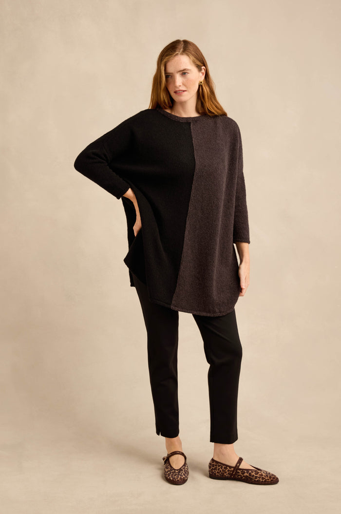 A woman stands posing in the Two Colour Knit, a wool-rich tunic featuring an asymmetric design in two-tone black and dark gray, paired with black pants and leopard print shoes against a beige background.