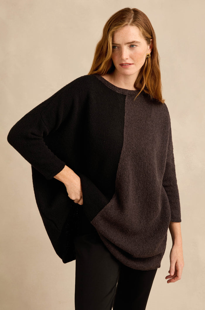 A woman is wearing the Two Colour Knit, a loose, long-sleeved sweater with an asymmetric colorblock design in black and dark gray, paired with black pants and standing against a neutral background.