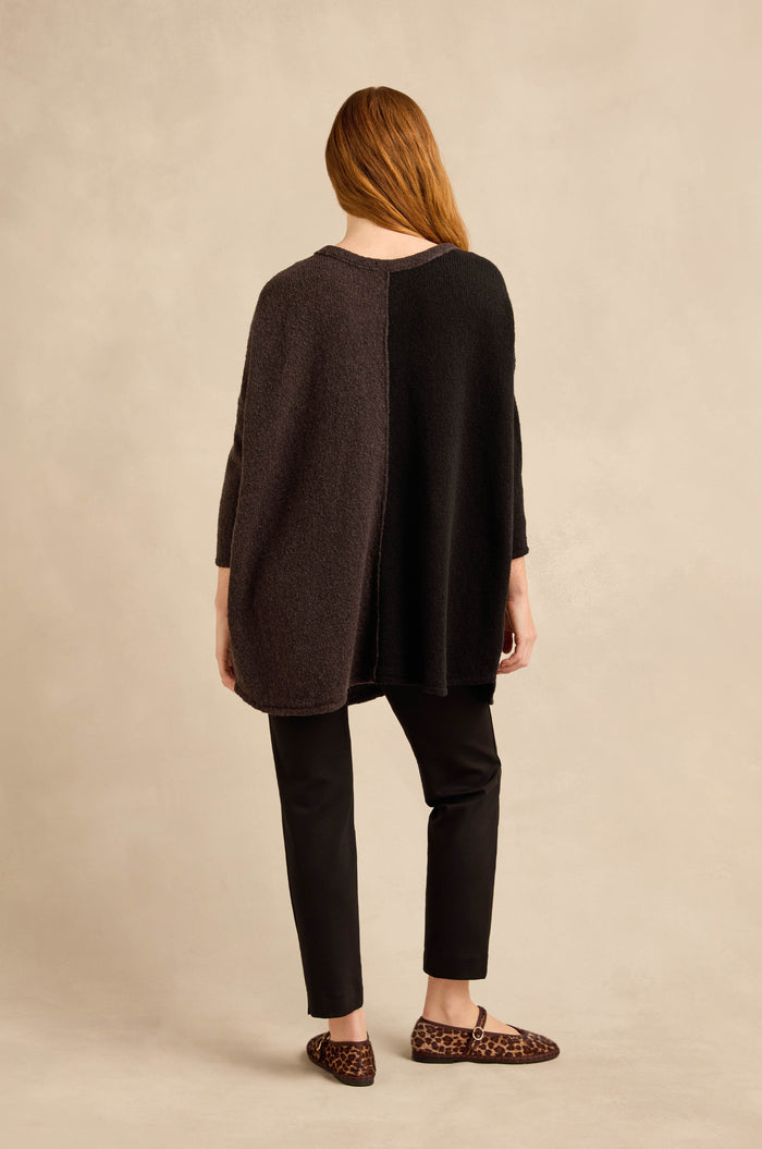 A person stands facing away from the camera, wearing the Two Colour Knit—an oversized sweater with an asymmetric design—paired with black pants and leopard-print loafers against a neutral background.