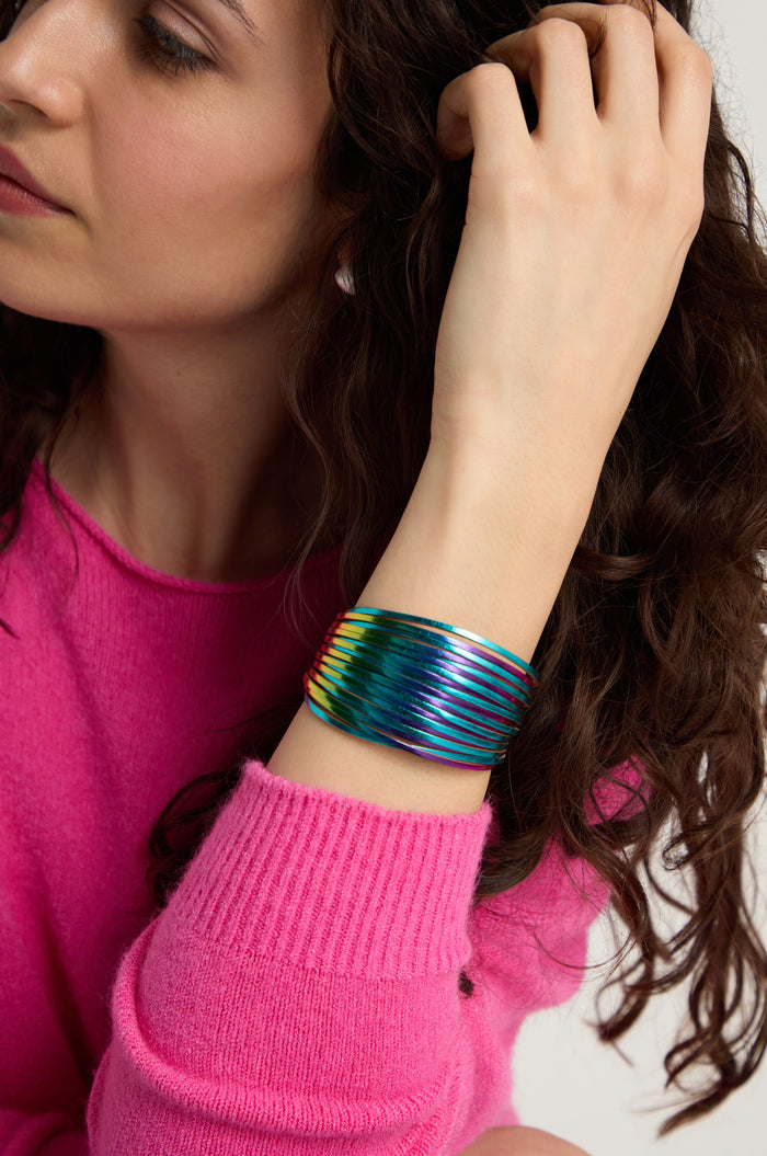 A person wearing a bright pink sweater shows off the Vegan Leather Metallic Bracelet on their wrist while touching their curly brown hair.