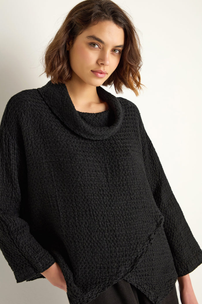 A woman with short brown hair wears the Waffle Linen Boxy Cowl Neck Top, a relaxed fit black textured sweater, standing against a plain light background.