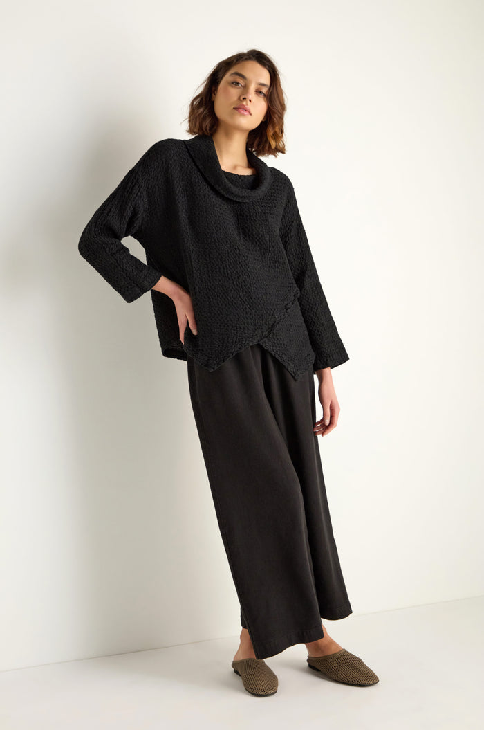 A woman stands against a plain background wearing the Waffle Linen Boxy Cowl Neck Top, relaxed black pants, and brown slip-on shoes.