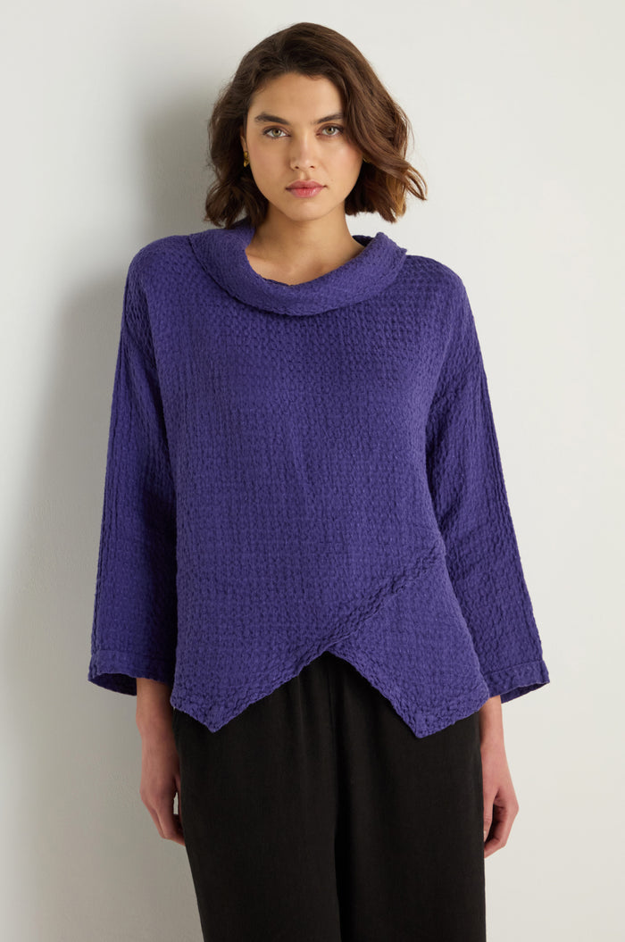 A woman stands against a plain light background wearing a relaxed fit, purple Waffle Linen Boxy Cowl Neck Top with long sleeves, an asymmetric hem, and black pants.