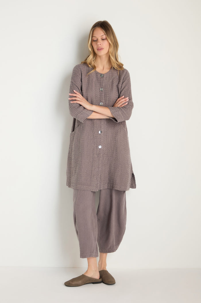 A woman stands against a plain background wearing the Waffle Linen Long Jacket in muted gray tones paired with matching pants and brown slip-on shoes—effortless, breathable wardrobe essentials.