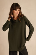 A woman stands against a plain beige background, looking downward, wearing the Waffle Textured Jersey Pine Cowl Neck Top—a long-sleeve, relaxed fit top in dark green with a distinctive waffle texture.