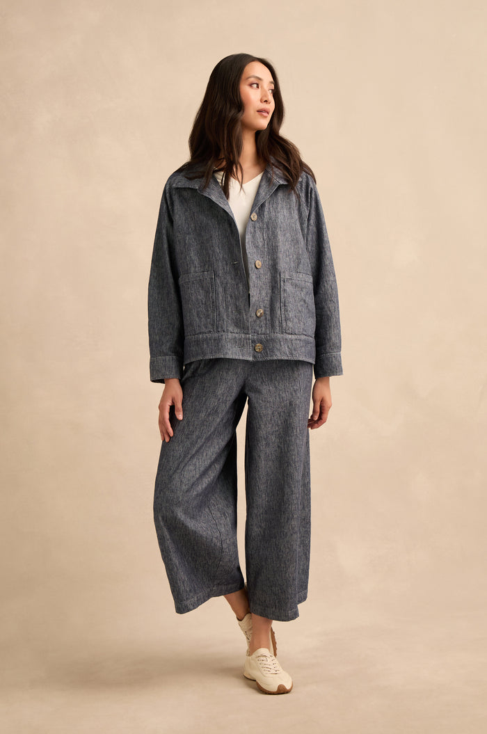 A woman stands against a plain background, wearing a loose-fitting Washed Cotton Linen Jacket and wide-leg pants with white sneakers, looking slightly to the side.