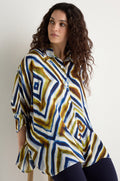 A woman with curly hair sits on a stool wearing the Wavy Lines Print Shirt, featuring geometric blue, green, and brown designs, paired with dark pants.
