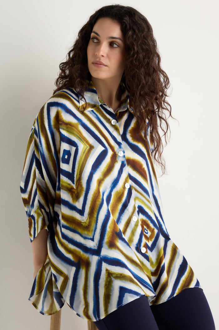 A woman with curly hair sits on a stool wearing the Wavy Lines Print Shirt, featuring geometric blue, green, and brown designs, paired with dark pants.