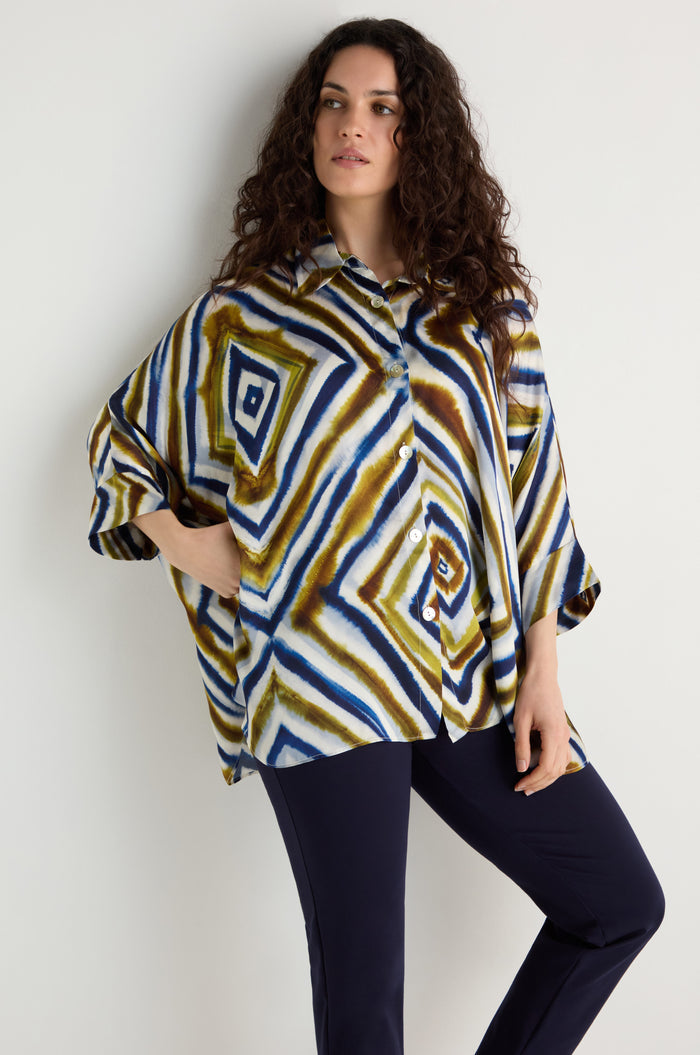 A woman with curly hair wears the Wavy Lines Print Shirt, a loose viscose top with blue, brown, and white abstract wavy patterns, paired with dark pants against a plain background.