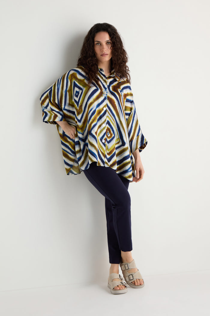 A woman stands against a white wall wearing the Wavy Lines Print Shirt, styled in a loose fit with dark pants and light sandals, one hand on her hip and the other by her side.
