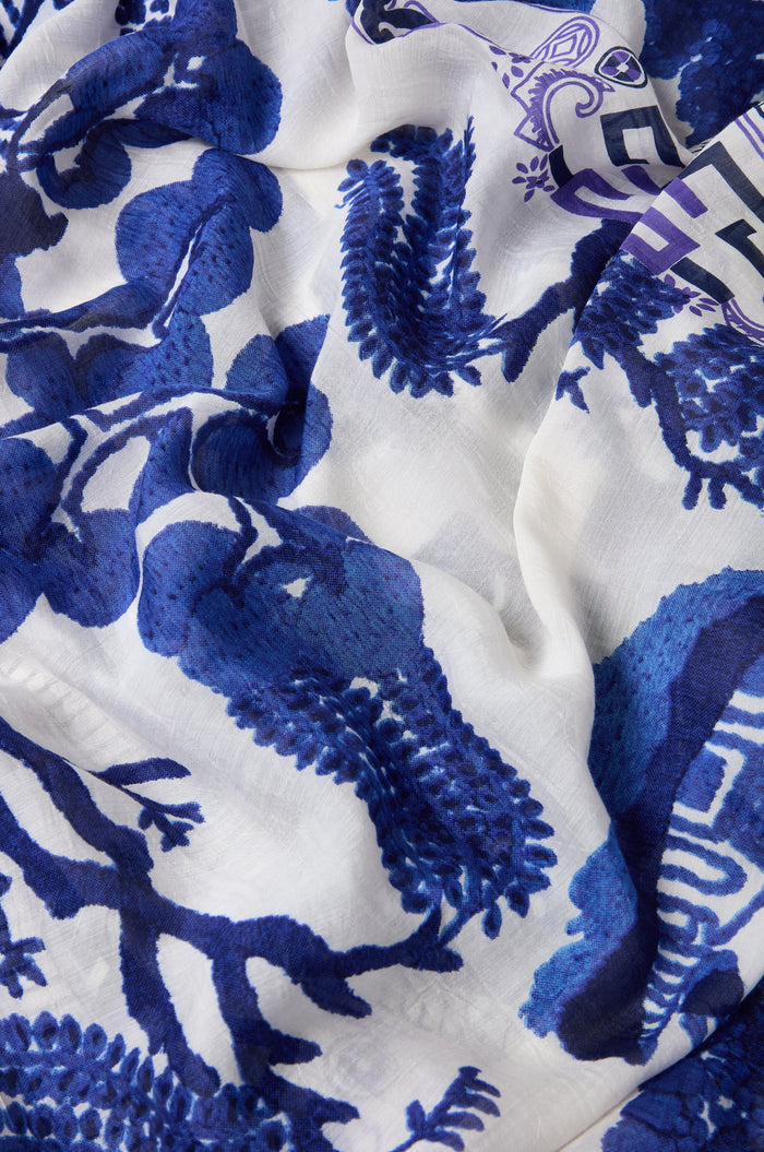The Willow Print Scarf features blue floral and leaf patterns on white fabric with intricate detailing and a slightly wrinkled texture, crafted from sustainable materials for effortless style.