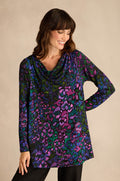 A woman stands against a plain background, wearing the Winter Garden Jersey Cowl Neck Top, which features long sleeves and a colorful abstract pattern in blue, green, purple, and black. With its flattering fit-and-flare silhouette and stylish cowl neck, she looks down with a slight smile.