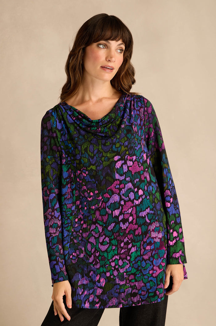 A woman wearing the Winter Garden Jersey Cowl Neck Top, featuring long sleeves and a multicolored leopard print with a draped cowl neckline, stands against a plain beige background.