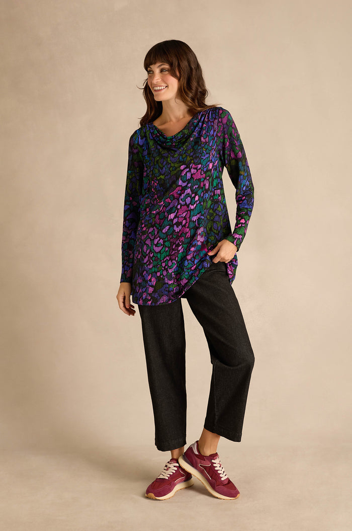 A woman stands in front of a neutral background, smiling with one hand in her pocket, wearing the Winter Garden Jersey Cowl Neck Top—featuring purple and blue patterns—paired with dark pants and maroon sneakers.