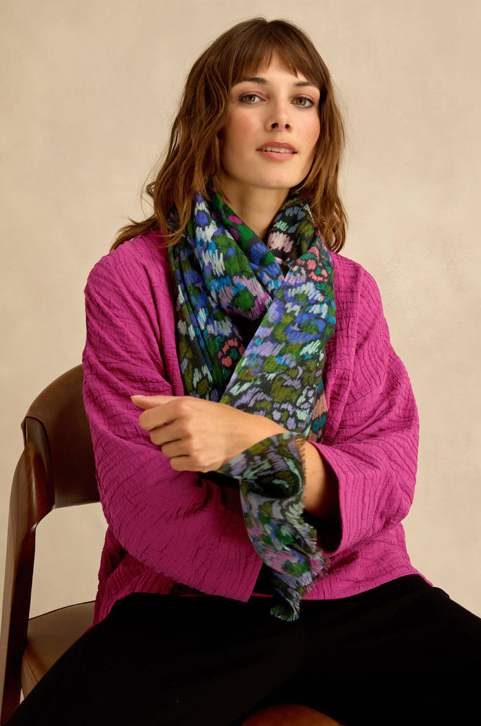 A woman sits on a chair against a neutral background, wearing a textured magenta jacket, black pants, and the Winter Garden Wool Scarf featuring a colorful botanical pattern.